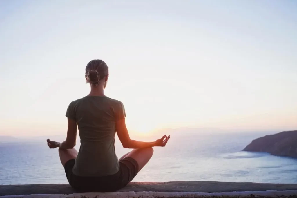 Woman sitting with prana mudra on a cliff in front of an ocean, meditating. Soft serene morning light.